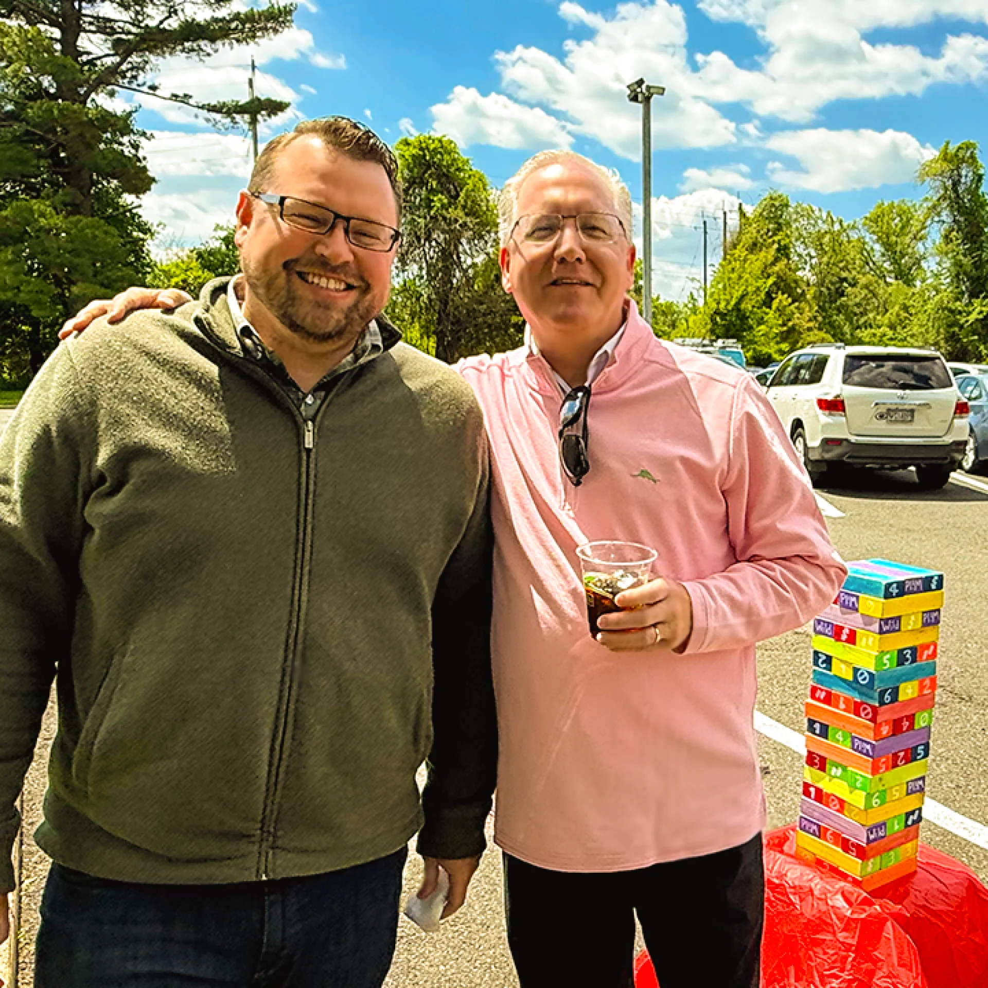 Our President of the company and an employee posing for a photo in front of the Jenga tower before round two. There is a blue sky with some clouds in the distance.