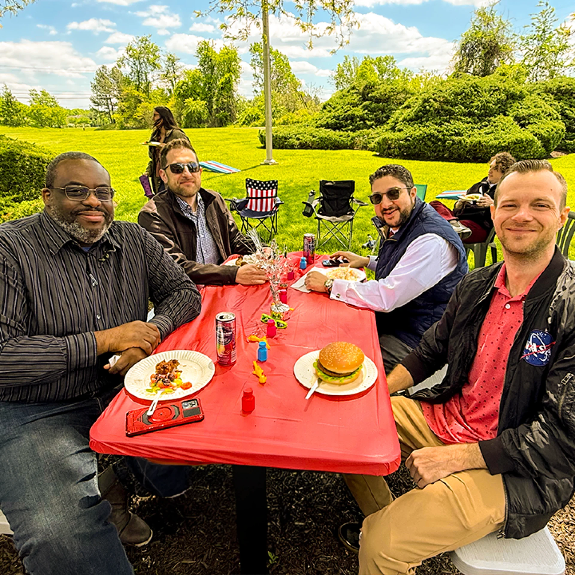 A group of leadership and employees eating together on a vibrant red tablecloth. The food on plates vary from burgers, coleslaw, to bbq. There are people in the background eating and walking around on the green grass.